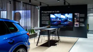 Bild eines Screens im Verkaufsraum eine Autohauses. Im Vordergrund sieht man einen blauen Audi dahinter eine Tischgruppe hinter der der Screen an einer Wand hängt